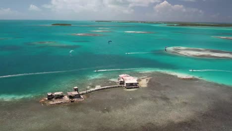 Stilt-houses-in-turquoise-sea-with-kite-surfers-around,-aerial-view