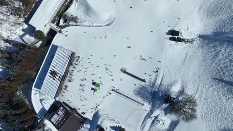 High-altitude-top-down-shot-of-bottom-of-ski-run,-ascending-shot-of-skiers-arriving-at-base-of-mountain-lining-up-for-the-chairlifts