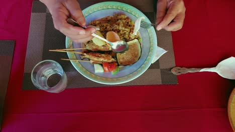 Overhead-view-of-eating-a-traditional-Indonesian-meal-with-nasi-goreng,-satay-skewers-served-with-saus-kacang-or-peanut-sauce-on-a-red-tablecloth