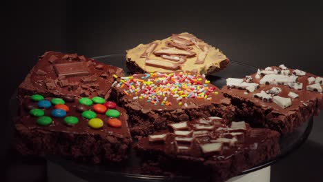 Vorderansicht-Eines-Tellers-Mit-Schokoladenbrownies-Auf-Einem-Drehteller,-Der-Sich-In-Einer-Schleife-Dreht,-Mit-Schwarzem-Hintergrund,-Mm-Hershey-Kinder-Oreo-Streusel,-Milchstraße,-Snickers-Kollektion