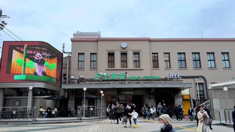 Crowded-entrance-of-a-historic-train-station-with-digital-billboard,-daytime