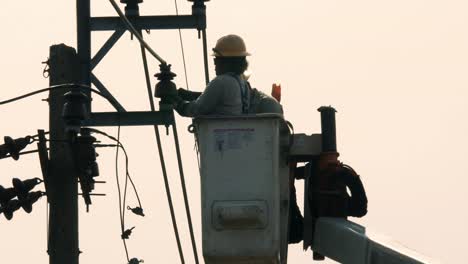 Putting-a-tape-around-a-terminal-of-an-electrical-post,-the-two-electric-repairmen-are-lifted-by-a-crane-to-fix-the-electrical-problem