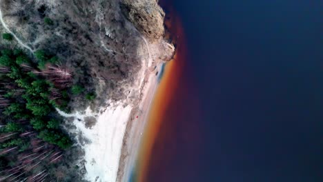 Aerial-Fly-Over-of-a-red-tidal-pool-along-a-rocky-coastline-with-green-trees-and-shrubs,-possibly-taken-with-a-drone