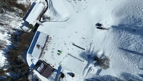 High-altitude-top-down-shot-of-bottom-of-ski-run,-static-shot-of-skiers-arriving-at-base-of-mountain-lining-up-for-the-chairlifts