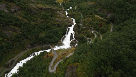 huge-waterfall-of-a-glacier-in-the-middle-of-a-green-forest,-people-hiking,-Briksdalsbreen,-norway,-nature,-drone