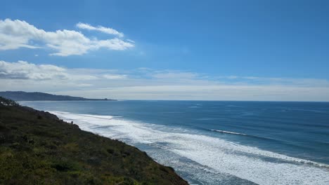 Scenic-panning-view-of-hiking-trail-in-Torrey-Pines,-San-Diego-facing-ocean-waves-crashing-against-shoreline-and-blue-skies