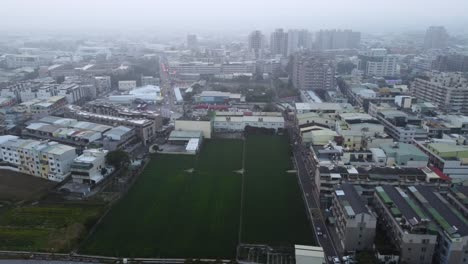 A-sprawling-urban-area-with-buildings-and-a-central-green-field-on-an-overcast-day,-misty-ambiance,-aerial-view