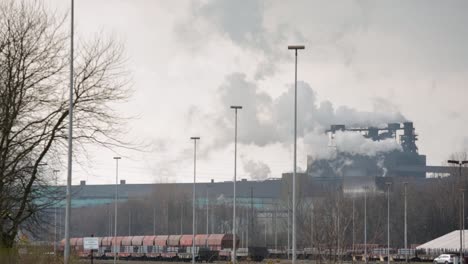 Industrial-plant-with-smokestacks-emitting-steam-against-a-cloudy-sky,-train-cargo-in-the-foreground
