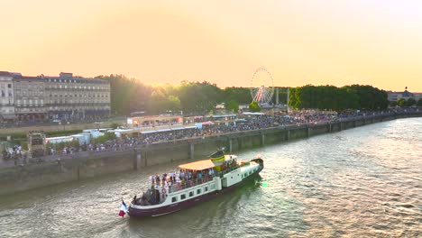 Gente-En-Un-Barco-Turístico-Navegando-Por-El-Río-Garona-Con-Noria-Y-Puesta-De-Sol-Durante-La-Feria-Del-Vino,-Toma-Aérea-A-La-Izquierda