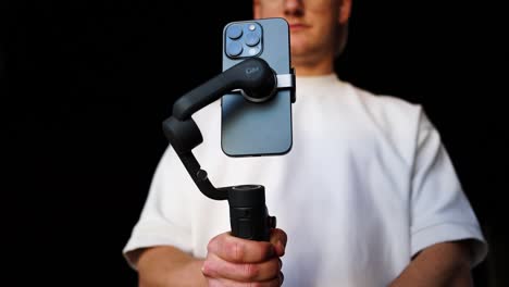 Male-stand-near-black-studio-background-and-turn-on-compact-mobile-stabilizer