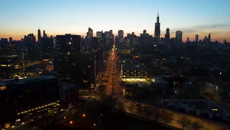 Eine-Faszinierende-Drohnen-Luftaufnahme-Von-Chicago-In-Der-Abenddämmerung-Nach-Links,-Die-Beleuchtete-Wolkenkratzer-Vor-Dem-Dunkler-Werdenden-Himmel-Zeigt.-Die-Goldenen-Farbtöne-Des-Sonnenuntergangs-Heben-Architektonische-Wunder-Hervor.