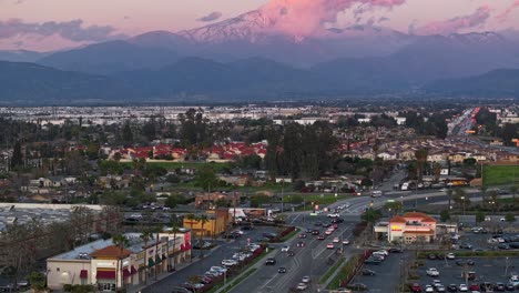 Timelapse-Sobre-Una-Concurrida-Intersección-En-San-Bernardino-California-Durante-La-Puesta-De-Sol-Con-Nubes-Rosadas-En-El-Fondo-Frente-A-Las-Montañas-Cubiertas-De-Nieve-Del-Bosque-Nacional-De-San-Bernardino-Estática-Aérea