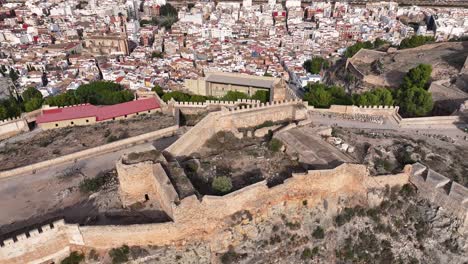 Vuelo-Con-Un-Dron-Realizando-Una-Rotación-De-Cámara-Visualizando-Ambas-Partes-De-Las-Murallas-De-La-Fortaleza-Y-La-Ciudad-De-Sagunto-Con-Sus-Casas-Al-Fondo-En-La-Mañana-En-Valencia-España