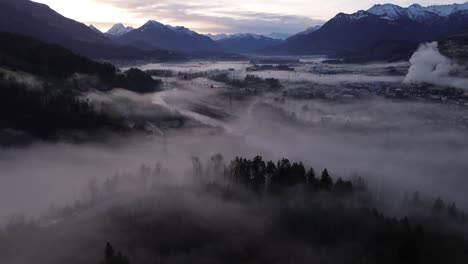 Aerial-Drone-shot-over-misty-Cityscape-at-Sunrise-Surrounded-by-Snow-Covered-Mountains-in-Frastanz,-Vorarlberg,-Austria