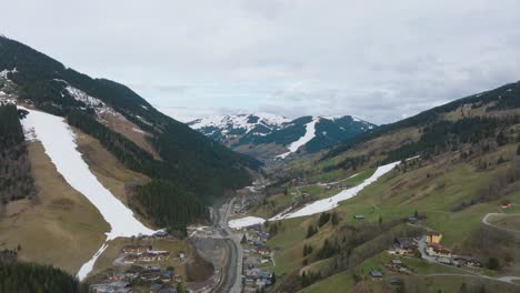 Saalbach-Hinterglemm-,-descending-towards-an-alpine-ski-resort-with-patchy-snow-in-spring,-aerial-view