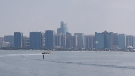 Abu-Dhabi,-Emiratos-Árabes-Unidos,-Frente-Al-Mar-Y-Horizonte-Urbano,-Lancha-Rápida-Y-Paracaídas-Parasailing,-Vista-Amplia