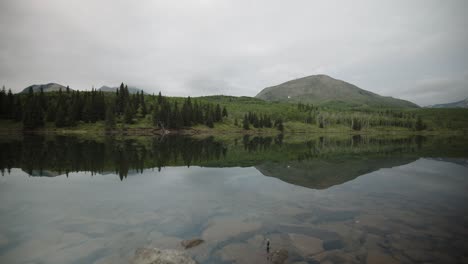 Un-Tranquilo-Espejo-De-Lago-De-Montaña-Refleja-El-Bosque-Circundante-Y-Las-Colinas-Onduladas-Bajo-Un-Suave-Cielo-Nublado,-Mostrando-La-Quietud-De-La-Naturaleza-Y-La-Naturaleza-Virgen