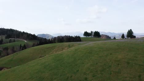 A-serene-expanse-of-lush-green-hills,-with-a-snaking-road-leading-to-a-secluded-farm,-all-set-before-a-dramatic-mountain-backdrop