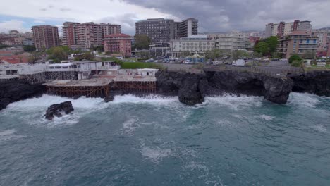 Aerial-low-altitude-shot-of-the-waterfront-of-Catania,-Sicily,-Italy-with-sea-waves-crashing-on-the-volcanic-cliff