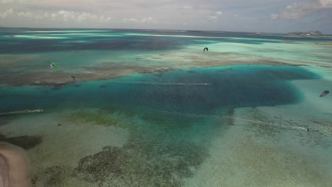 A-vibrant-cay-with-kite-surfers-on-clear-waters,-sunny-tropical-scene,-aerial-view