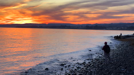 People-fishing-by-the-beach-with-an-incredible-orange-sunset-sky-in-Marbella-Estepona-Spain,-amazing-view,-calm-sea-water-waves,-4K-shot