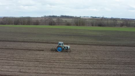 Aerial-view-from-side-of-old-blue-tractor-cultivate-crop-field,-Latvia
