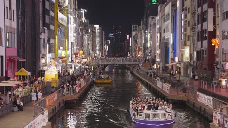 Toma-Nocturna-De-Barcos-Pasando,-Personas-Y-Letreros-Iluminados-Vista-Desde-El-Puente-Ebisu-Canal-Dotonbori-Namba-Osaka,-Japón