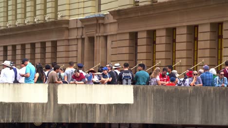 Familias-Australianas-Alineadas-En-El-Puente-Aéreo-Sobre-La-Calle-Adelaida,-Esperando-El-Comienzo-De-La-Tradición-Anual-Del-Desfile-Del-Día-De-Anzac,-Observando-El-Desfile.