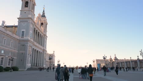 Turistas-Y-Lugareños-Caminan-Por-La-Plaza-Armería-Frente-A-La-Catedral-Católica-Romana-De-La-Almudena,-Terminada-En-1993,-Mientras-El-Atardecer-Comienza-A-Asentarse-En-Madrid,-España.