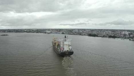 Cargo-ship-sailing-in-the-Suriname-river-towards-harbor,-drone-view,-city-in-background