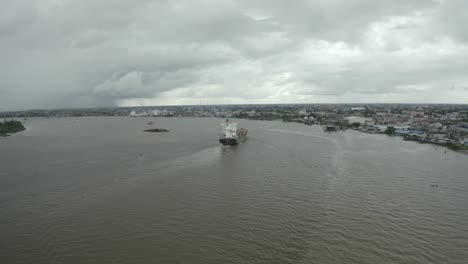 Cargo-ship-sailing-in-the-Suriname-river-towards-harbor,-drone-view-dolly-out,-city-in-background