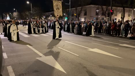 La-Procesión-Va-En-Formación-Donde-Vemos-El-Estandarte-De-La-Cofradía-De-La-Virgen-De-Los-Dolores-Detrás-De-Los-Nazarenos-Con-Niños-Y-Después-De-Las-Monjas-De-Negro-Vemos-Gente-A-Los-Lados