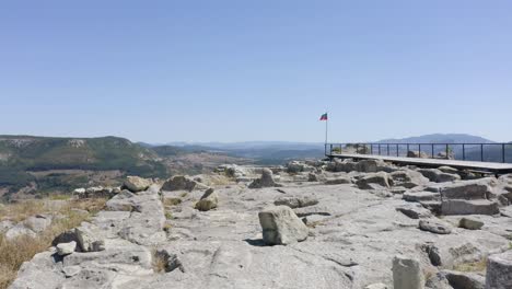 Approaching-drone-shot-showing-a-flag-being-blown-by-the-wind-at-the-end-of-a-viewpoint-platform-on-the-ancient-historical-site-of-of-Perperikon,-located-in-the-province-of-Kardzhali,-in-Bulgaria