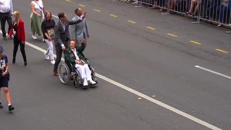 Senior-woman-veteran-in-a-wheelchair,-being-pushed-down-the-street-by-her-family,-proudly-waving-at-the-cheering-crowds-during-the-Anzac-Day-parade-in-Brisbane-city