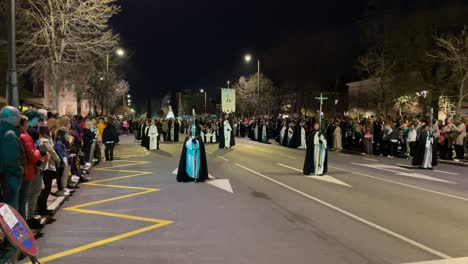 La-Procesión-Recorre-Una-Calle-Con-Formación-Donde-Vemos-En-Primer-Plano-A-Tres-Feligreses,-Uno-Con-Una-Cruz-Y-Dos-Con-Velas-Detrás,-Uno-Portando-Un-Estandarte,-Hacen-Una-Parada