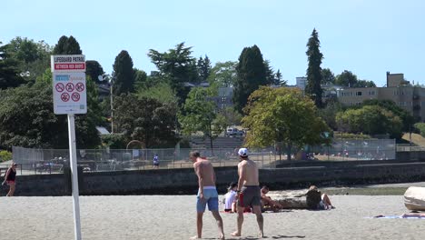 Ein-Mann-Ohne-Hemd,-Der-An-Einem-Strand-Mit-Sonnenanbetern-Und-Mehreren-Wanderern-Und-Bergen-Spazieren-Geht