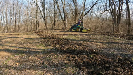Man-driving-a-utility-tractor-to-rototill-soil-in-a-deer-food-plot-between-timber-and-field-in-early-spring-in-Midwest