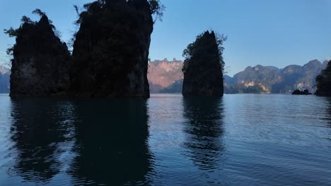 Vista-De-Las-Rocas-En-El-Parque-Nacional-Khao-Sok-De-Tailandia-Reflejándose-En-La-Superficie-Del-Agua-Con-Montañas-Tropicales-Y-Bosques-Al-Fondo