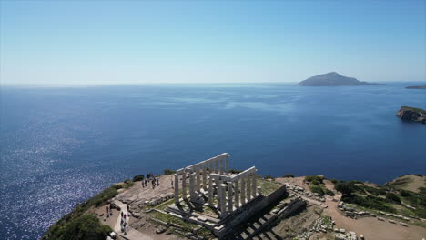 Aerial-drone-view-of-the-Temple-of-Poseidon-ruins-at-Sounion,-Greece,-with-visitors-and-a-distant-island