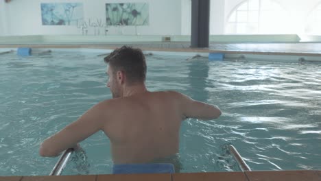 A-white-man-swims-in-an-indoor-pool-and-leans-on-handrails