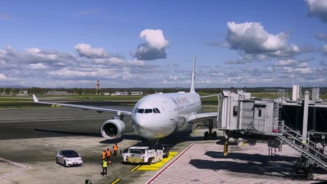 Camión-Pushback-Empujando-Aviones-De-Air-Canada-Hacia-Atrás-Para-Salir-De-La-Puerta-De-La-Terminal-Del-Aeropuerto-Con-Un-Puente-De-Carga-Separado-Y-Operadores-Terrestres-Trabajando-En-El-Asfalto