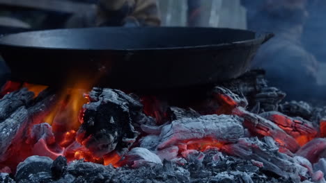 Large-cast-iron-skillet-placed-onto-hot-orange-bonfire-embers-in-camp