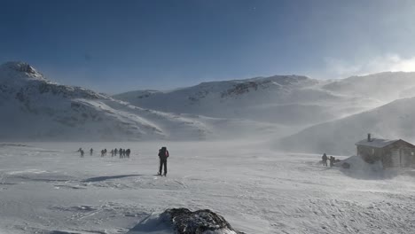 Gruppe-Von-Menschen-In-Windigen-Bergen-Zu-Ostern---Schnee-Weht-In-Zeitlupe-Um-Menschen-Und-Hütten-Herum