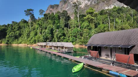 The-vibrant-shoreline-scenery-of-Khao-Sok-National-Park-in-Surat-Thani,-Thailand,-features-a-coastal-floating-hut-against-a-lush-backdrop-of-greenery-and-majestic-mountains