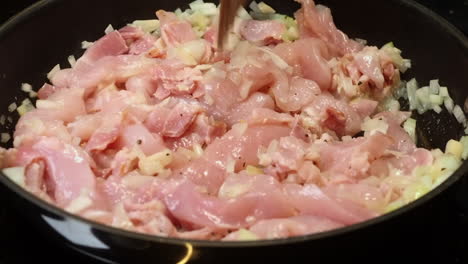 Raw-chicken-breast-added-to-frying-onions-in-hot-pan-on-stove,-closeup