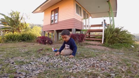 Niño-Jugando-Con-Piedras-Sentado-En-El-Suelo,-Frente-A-Una-Pequeña-Casa-De-Campo-Ubicada-En-La-Plantación-En-La-Cima-De-Una-Colina-En-Phetachabun,-Tailandia