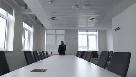 Male-Sat-And-End-Of-Long-Boardroom-Table-In-Empty-Conference-Room-Speaking-On-Mobile-Phone