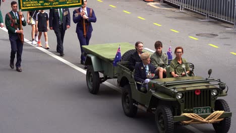 Veteran-and-families-ride-in-a-vintage-military-vehicle-with-a-trailer,-driving-down-the-street,-participating-in-the-Anzac-Day-parade-in-Brisbane-city