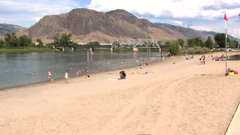 River-beach-with-lady-walking,-child-running,-sunny-day-sunbathers