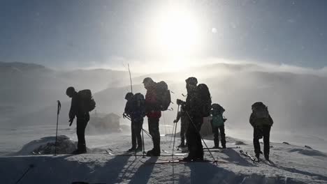 Grupo-De-Personas-Paradas-En-Una-Montaña-Nevada-Durante-Una-Caminata-Ventosa,-Silueta-Frente-Al-Sol,-Cámara-Lenta